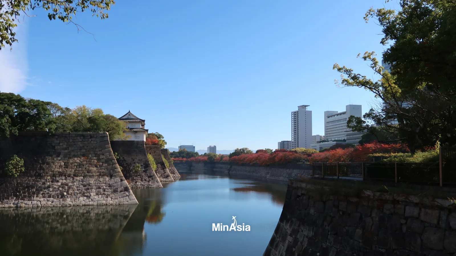 Paisagem serena do parque do castelo de Osaka, com visitantes passeando e a marca MinAsia Tours discretamente exibida.