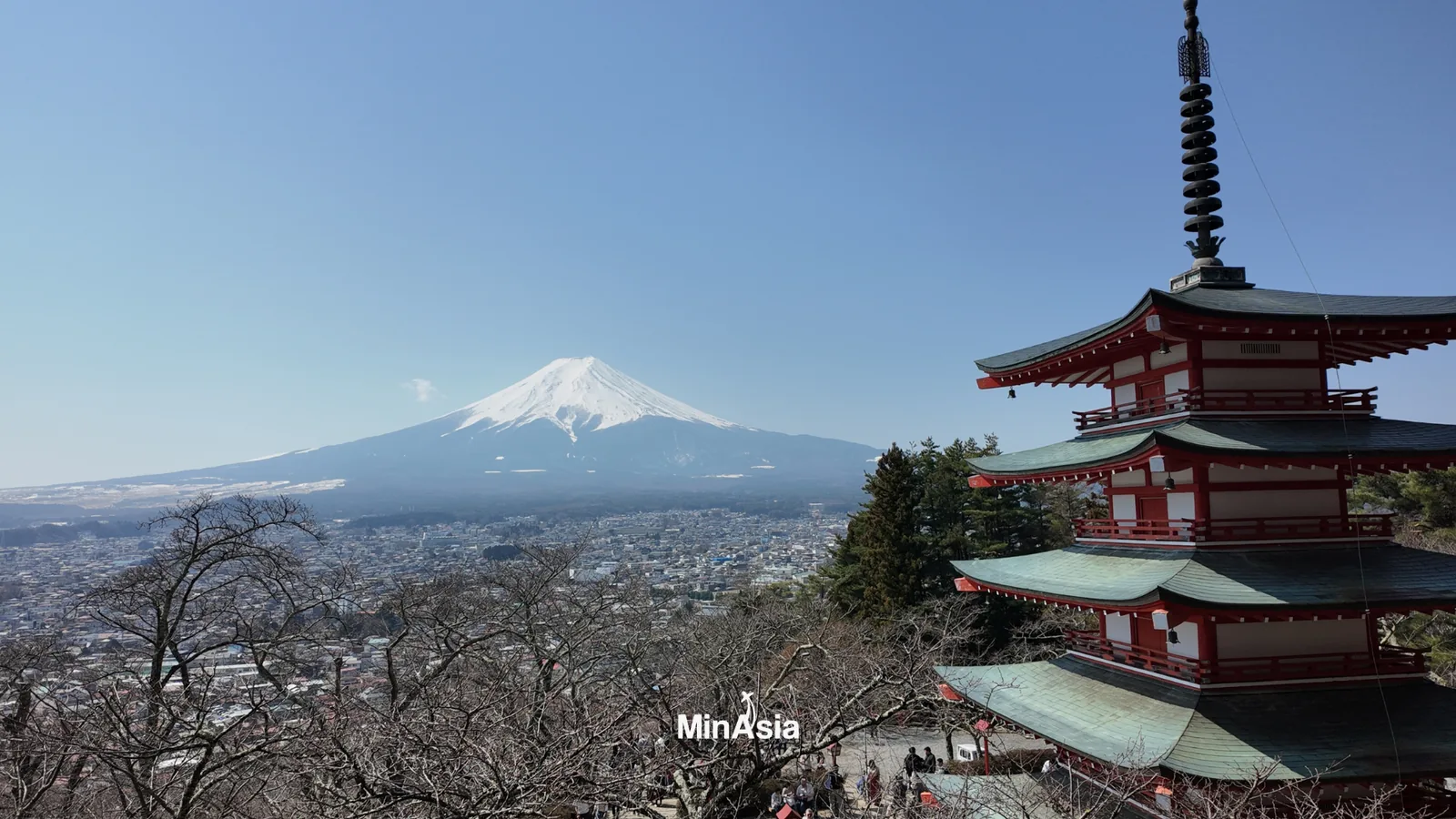 Um viajante admirando o monte fuji da pagoda Chureito em um dia ensolarado. A estrutura vermelha contrasta com o azul do céu e a vegetação exuberante.